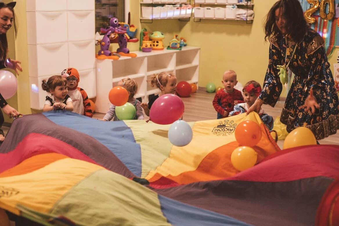 Bal karnawałowy w żłobku - dzieci bawią się na balu przy chuście animacyjnej i latających balonach.